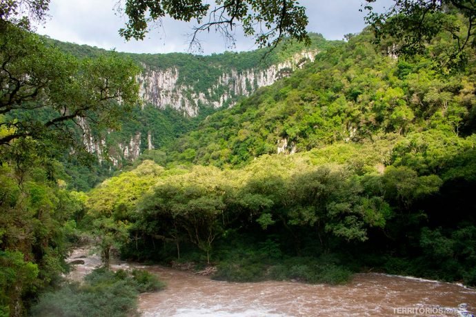 Rio com águas turvas e paredão de pedra com muito verde em Boqueirão do Leão, um dos destaques do Caminho das Cascatas